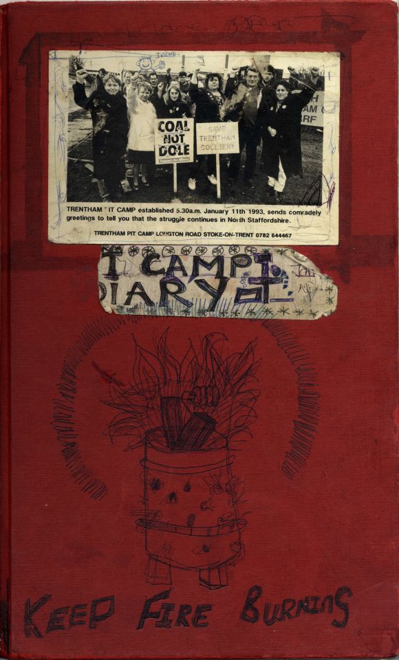 Front cover of a red diary with a partially torn label that reads 'T Camp Diary,' likely originally saying 'Pit Camp Diary.' The cover features a sketch of a makeshift fire in a garden incinerator bin with the words 'Keep Fire Burning.’ A newspaper clipping is also attached, showing the founding members rallying for support in 1993.